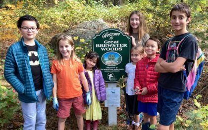 Seven children of varying ages standing in the woods in front of a sign that reads in part: "Trail maintenance by the youth of First Parish Church."