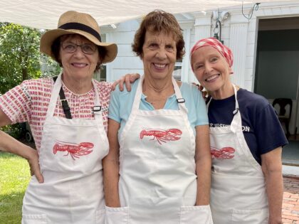 Three volunteers wearing white aprons with lobsters on them standing under a white canopy outside of a white house