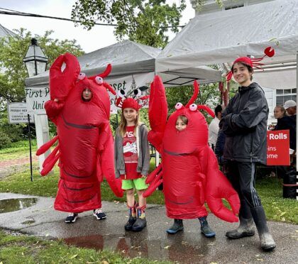 Four young people, two wearing red lobster mascot costumes and two wering lobster head gear, standing outside in the rain to gather attention for lobster roll customers.