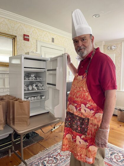 Man with apron and chef's hat standing in front of open refrigerator stacked with foil wrapped lobster rolls.