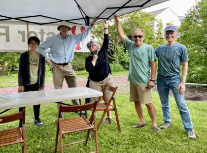 Five people standing outside on grass under a white canopy tent holding it up to be secured. Also shown are a rectangular table and two lawn chairs.