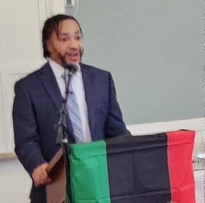 Guest Speaker, De'Shawn Washington, first black Massachusetts Teacher of the year, at podium draped with Pan-African flag - black, red, and green striped.