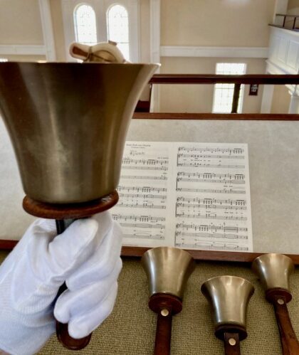cloth gloved hand shown holding a gold/brass bell with three additional bells on a table in front of sheet music.