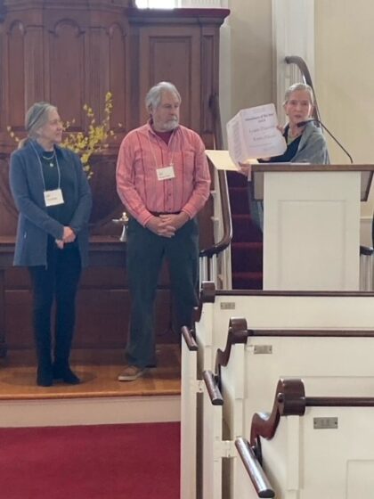 Three people shown on Meetinghouse alter. Speaker is at the pulpit, honorees are to the left listening to the speaker as the award certificates are shown.