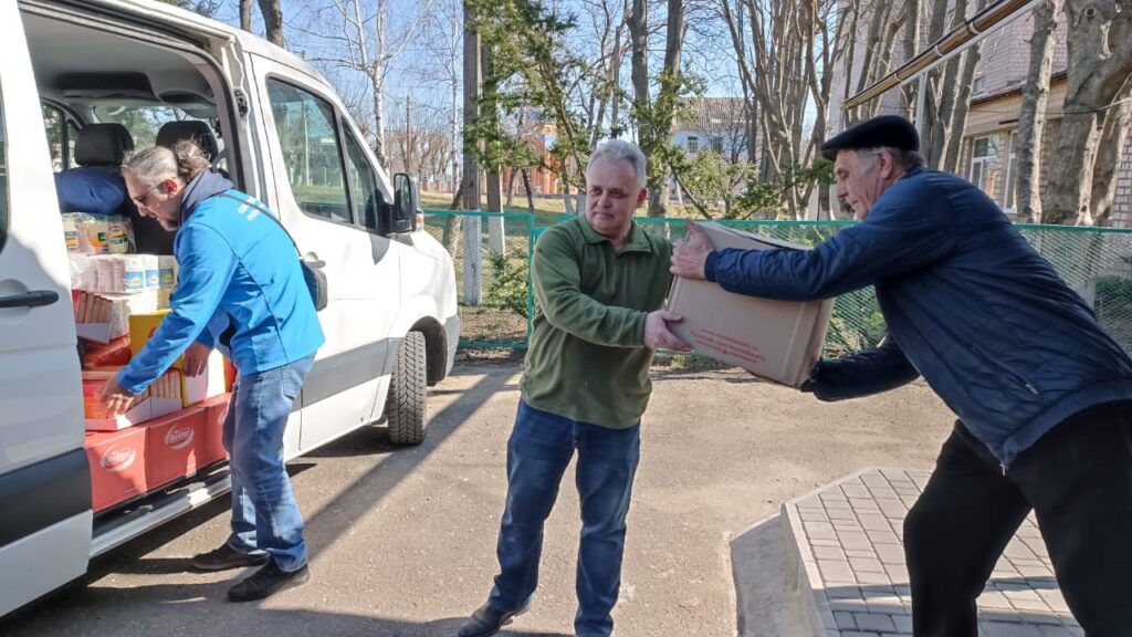 A van, with side door open to reveal a large quantity of supplies, is being unpacked by a team (three members shown) handing one package to the other.