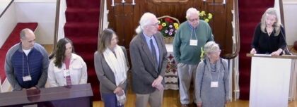 Six people standing on an alter facing right towards one speaker at the podium.